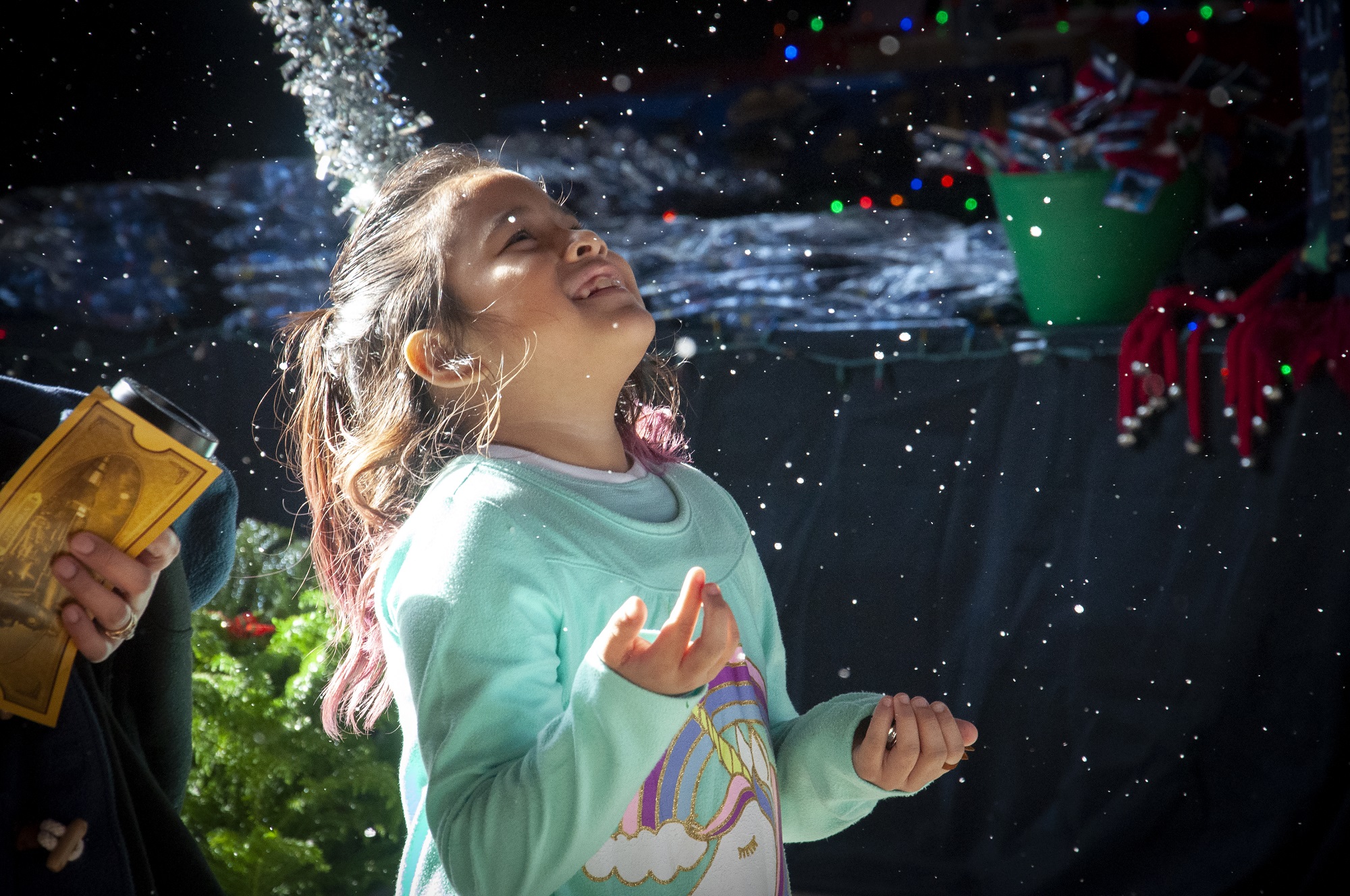 This is an image of a girl standing in the snow with her hands. There is a golden ticket in someone's hands in the background.